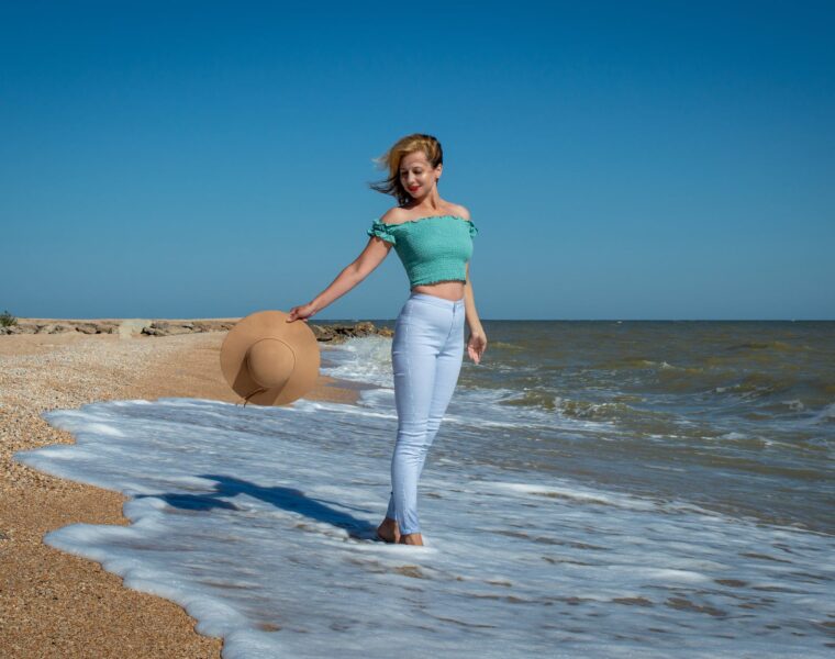 Poruke Svemira Naučite biti svoj broj 1., a woman in teal tank top and blue pants holding a brown hat standing on the beach