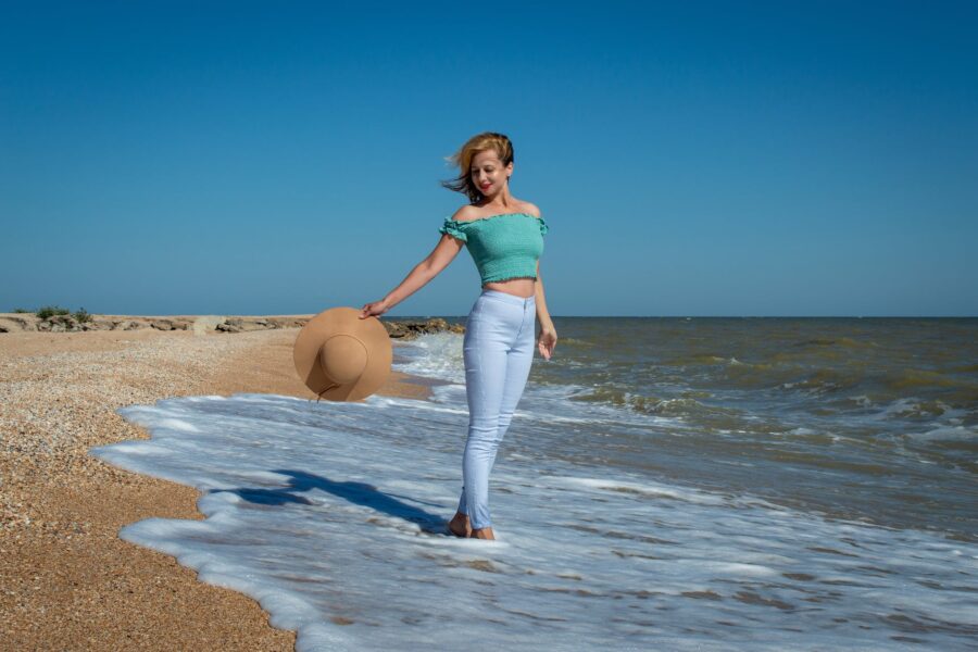 Poruke Svemira Naučite biti svoj broj 1., a woman in teal tank top and blue pants holding a brown hat standing on the beach